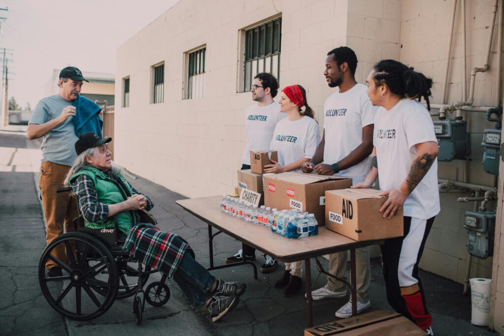 Projetos Volunteers assist a senior adult in a wheelchair by distributing aid packages outdoors.